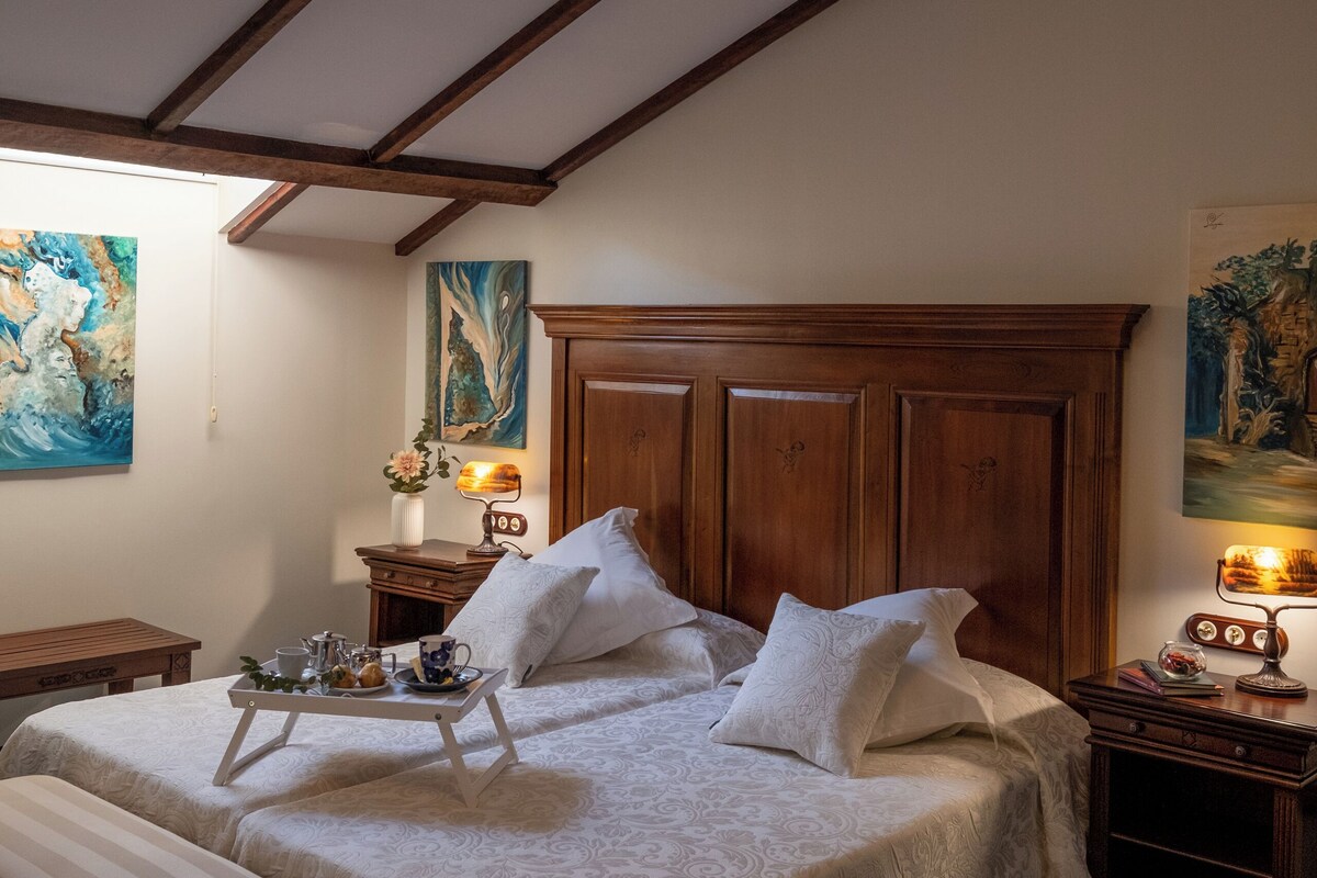 Une chambre avec un plafond en bois, un lit recouvert d’une courtepointe blanche, un banc, une table de chevet et une lampe fixée au mur.
