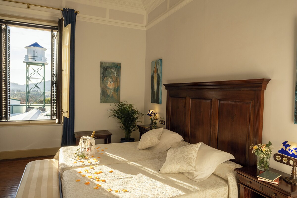 Une chambre bien éclairée, avec un grand lit, une table de chevet avec une lampe et une vue sur une tour par une fenêtre ouverte.