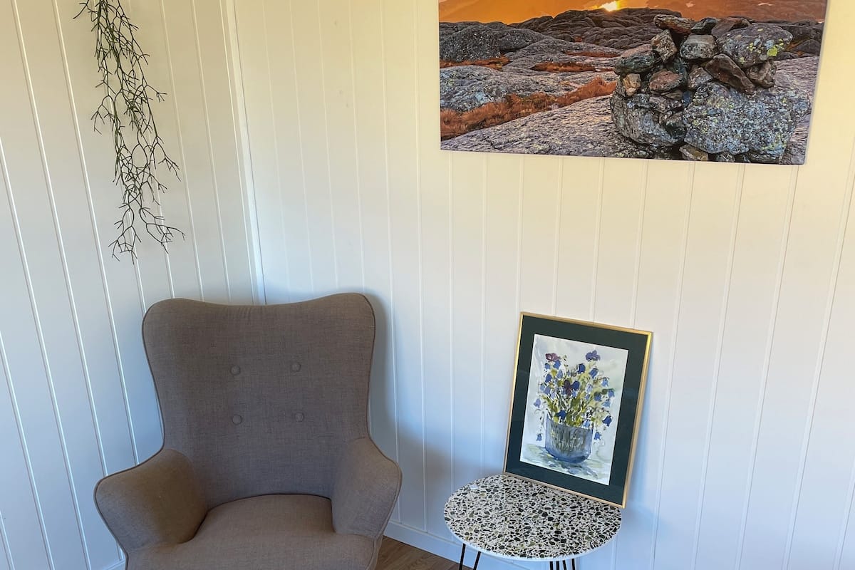 A room with a grey armchair, a small round table with a framed picture, and a wall-mounted plant.
