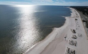 Ovenfra set er der en strand med klart himmel, sandet bredde og en vej der løber parallelt med stranden.