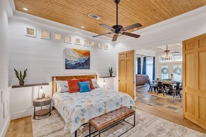 Um quarto com cama, ventilador de teto, uma pintura grande, área de jantar com mesa e cadeiras, e uma porta de madeira.