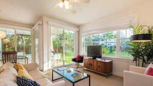 A spacious living room with a glass coffee table, white sofas, and a television on a wooden cabinet. - Light and Airy North Shore Condo. Owner hosted w/on site Co-host!  (Princeville)