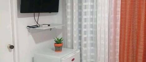 A room with a flat-screen TV mounted on the wall, a small white cabinet with a potted plant, and a door with a handle.