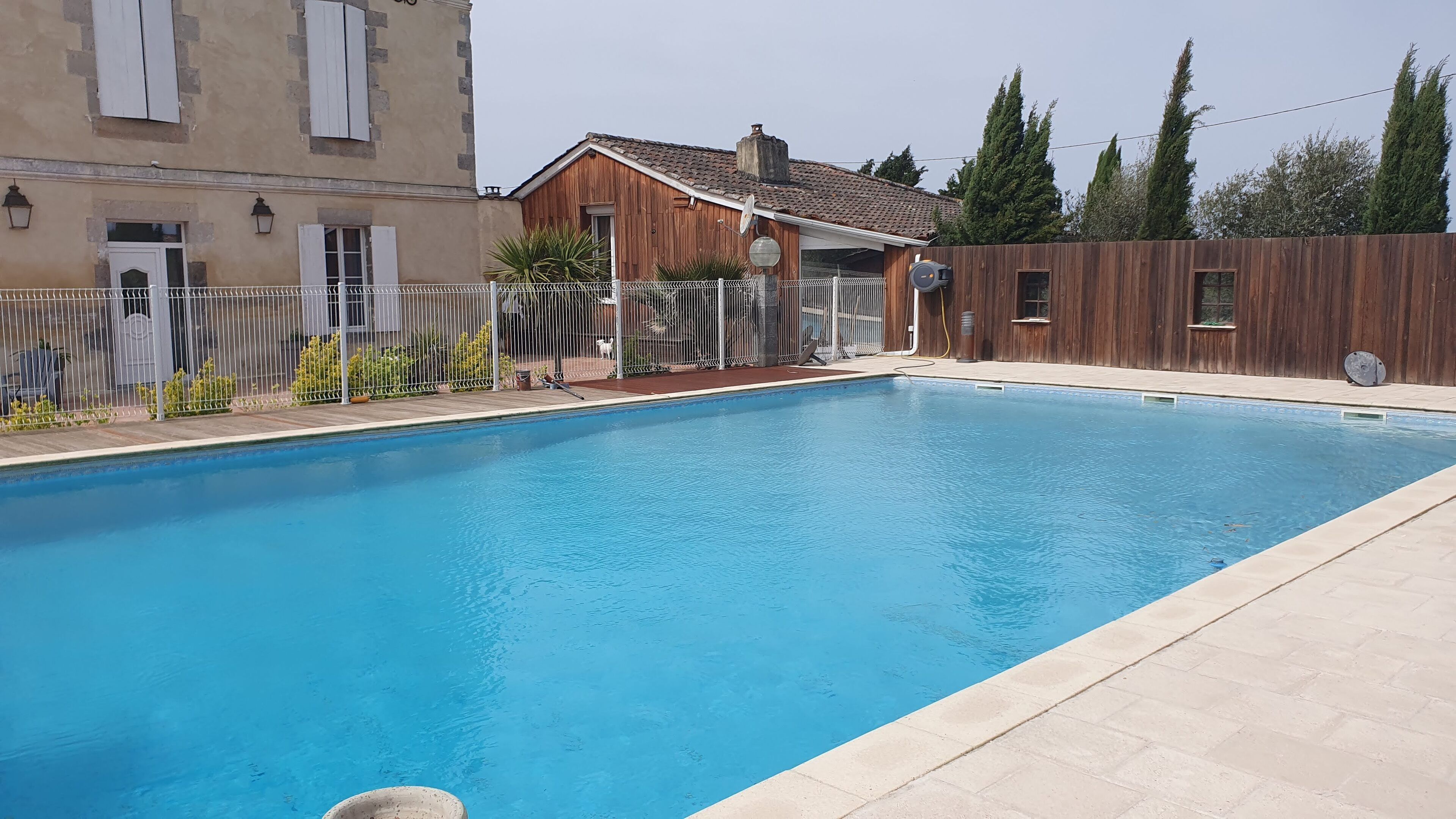 Une piscine rectangulaire entourée d’une clôture en bois, avec un bâtiment doté d’un toit en tuiles en arrière-plan.