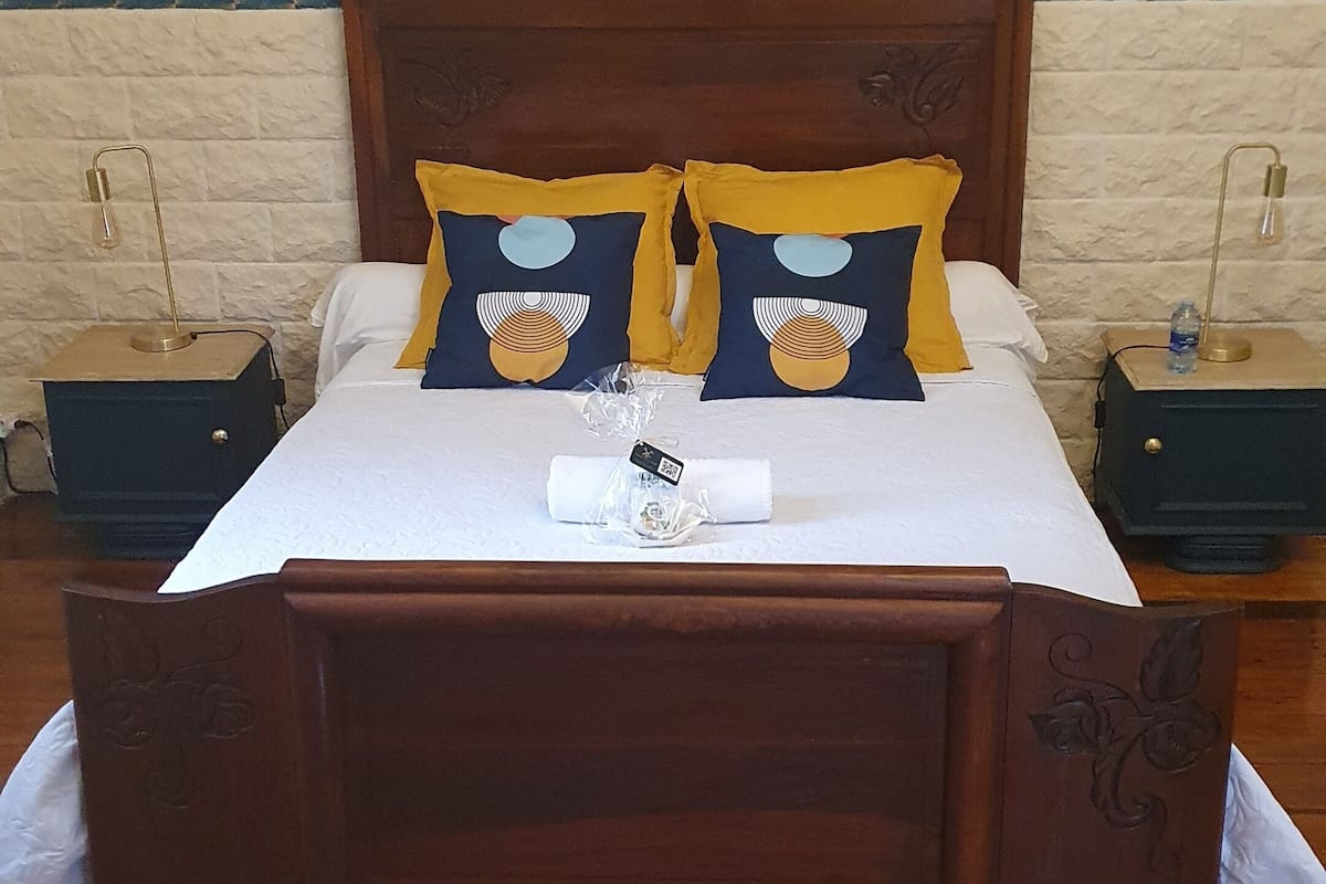 Uma cama de madeira com cabeceira, dois travesseiros com desenho de sol, lençol branco e uma pequena mesa de cabeceira com abajur.