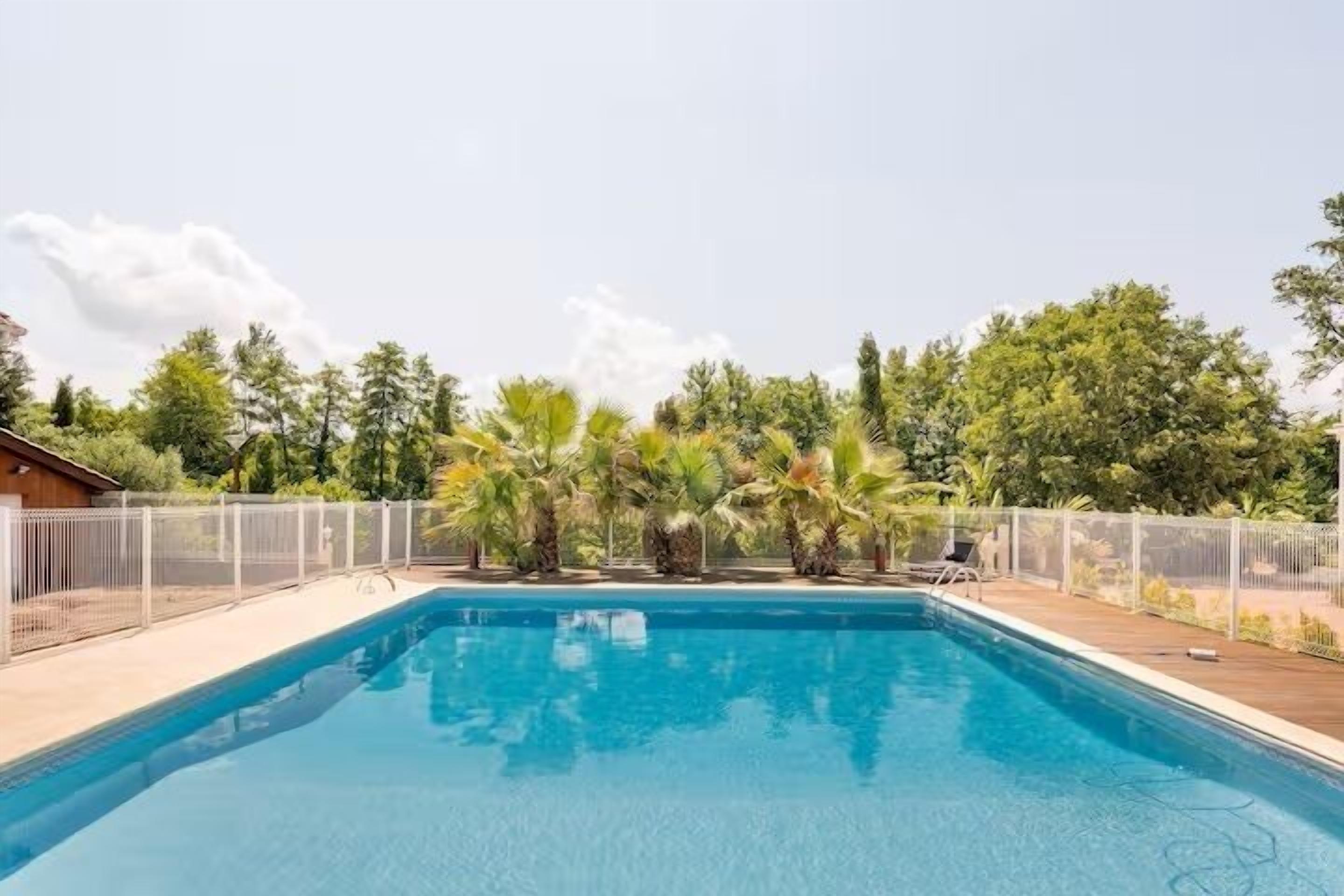 Una piscina rectangular con cubierta de madera, rodeada por una valla blanca y palmeras.