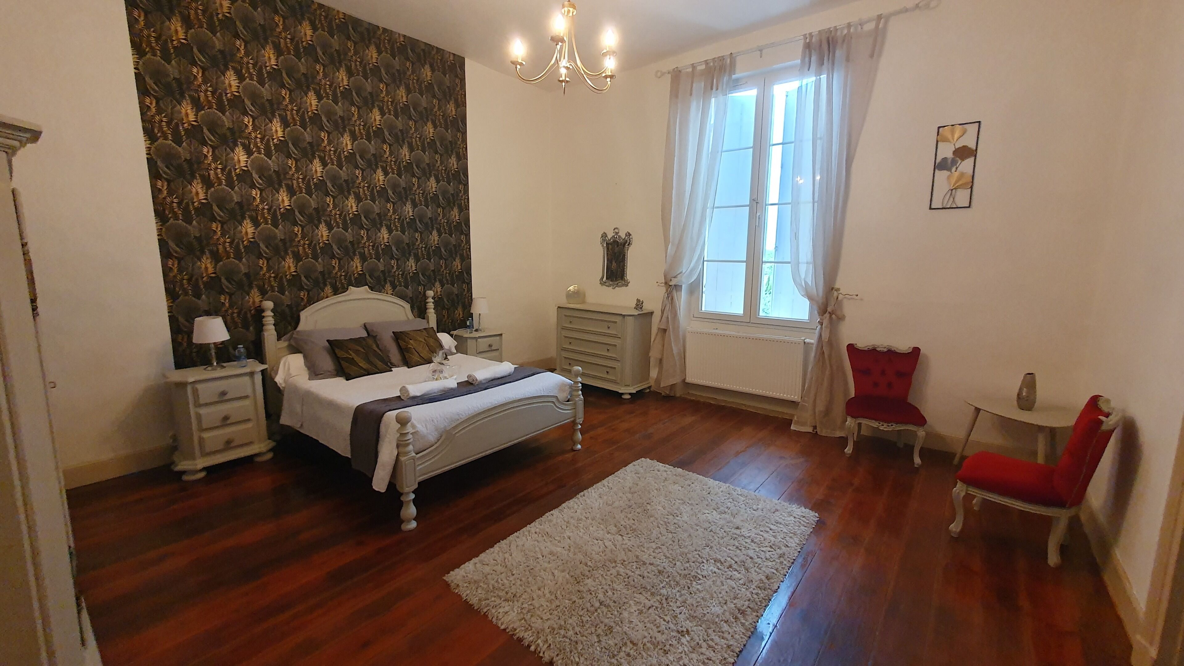 A bedroom with a bed, bedside tables, a dresser, a chair, a window with curtains, and a chandelier.