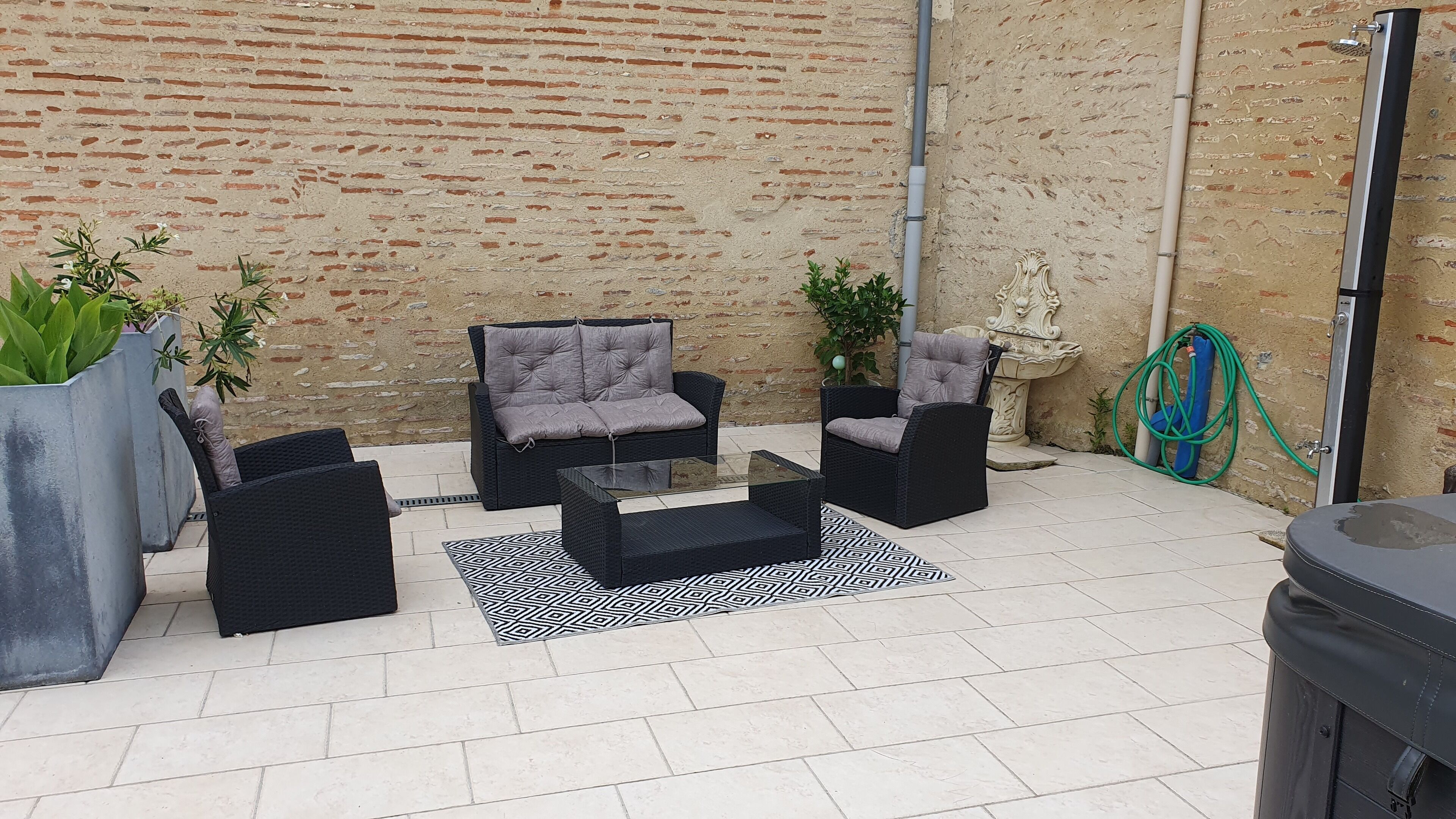Un patio al aire libre con mobiliario de mimbre negro, incluyendo dos sofás y una mesa de centro, sobre una alfombra estampada. Se ven plantas en macetas y una fuente decorativa al fondo.