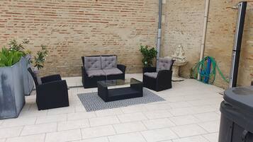 Un patio al aire libre con mobiliario de mimbre negro, incluyendo dos sofás y una mesa de centro, sobre una alfombra estampada. Se ven plantas en macetas y una fuente decorativa al fondo.