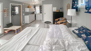 Ein Schlafzimmer mit einem großen Bett, einem Schreibtisch und einem Stuhl. Ein kleiner Tisch mit einem Laptop und einem Korb steht dort. Das Zimmer hat eine Tür zu einer Küchenzeile.