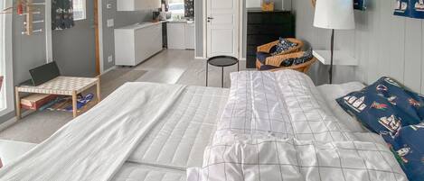 A bedroom with a large bed, a desk, and a chair. There is a small table with a laptop and a basket. The room has a door leading to a kitchen area.