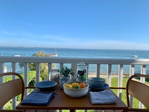 Ein Tisch mit einer Obstschale, einem mit 'TEE' beschrifteten Einmachglas und einer blauen Schüssel auf einem Balkon mit Blick aufs Meer.