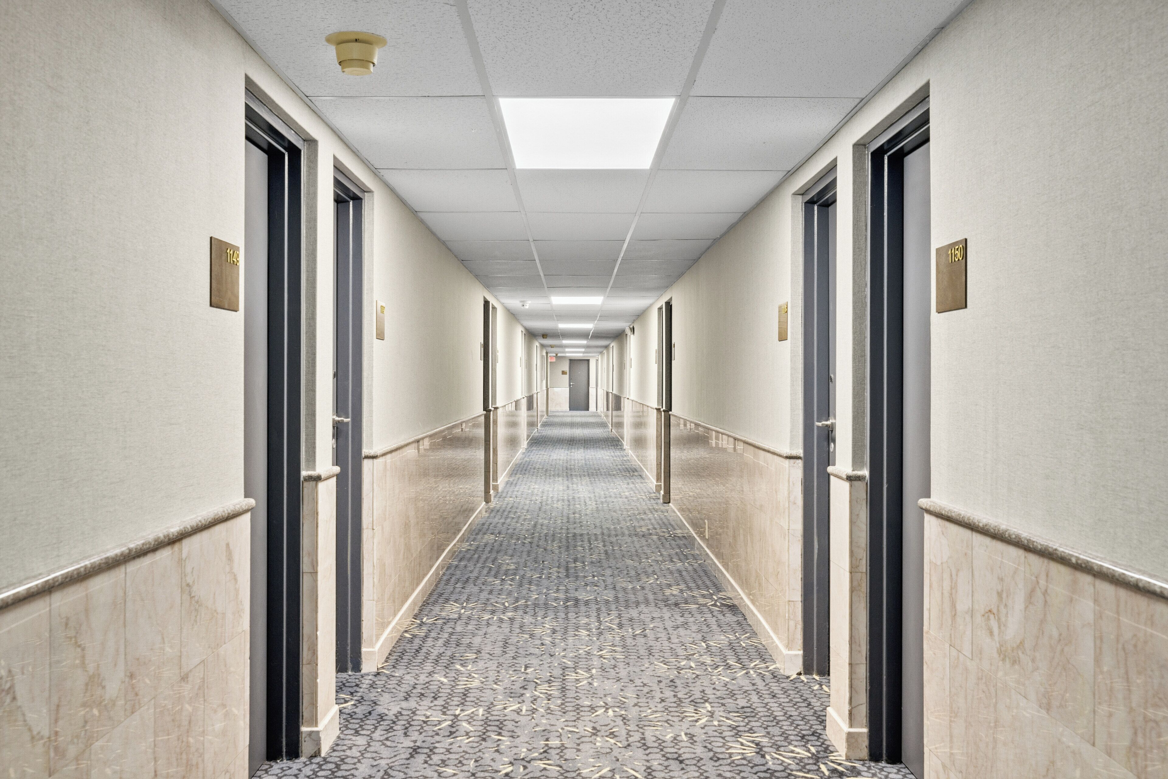Passadís d'hotel amb una catifa estampada, portes numerades i una làmpada al sostre.