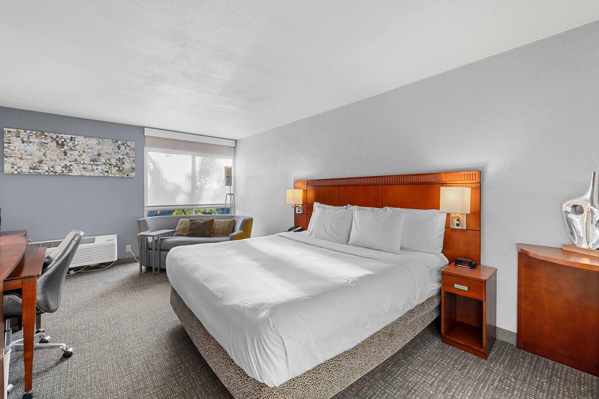 Quarto de hotel com uma cama grande, uma escrivaninha com cadeira, uma mesinha e uma luminária.