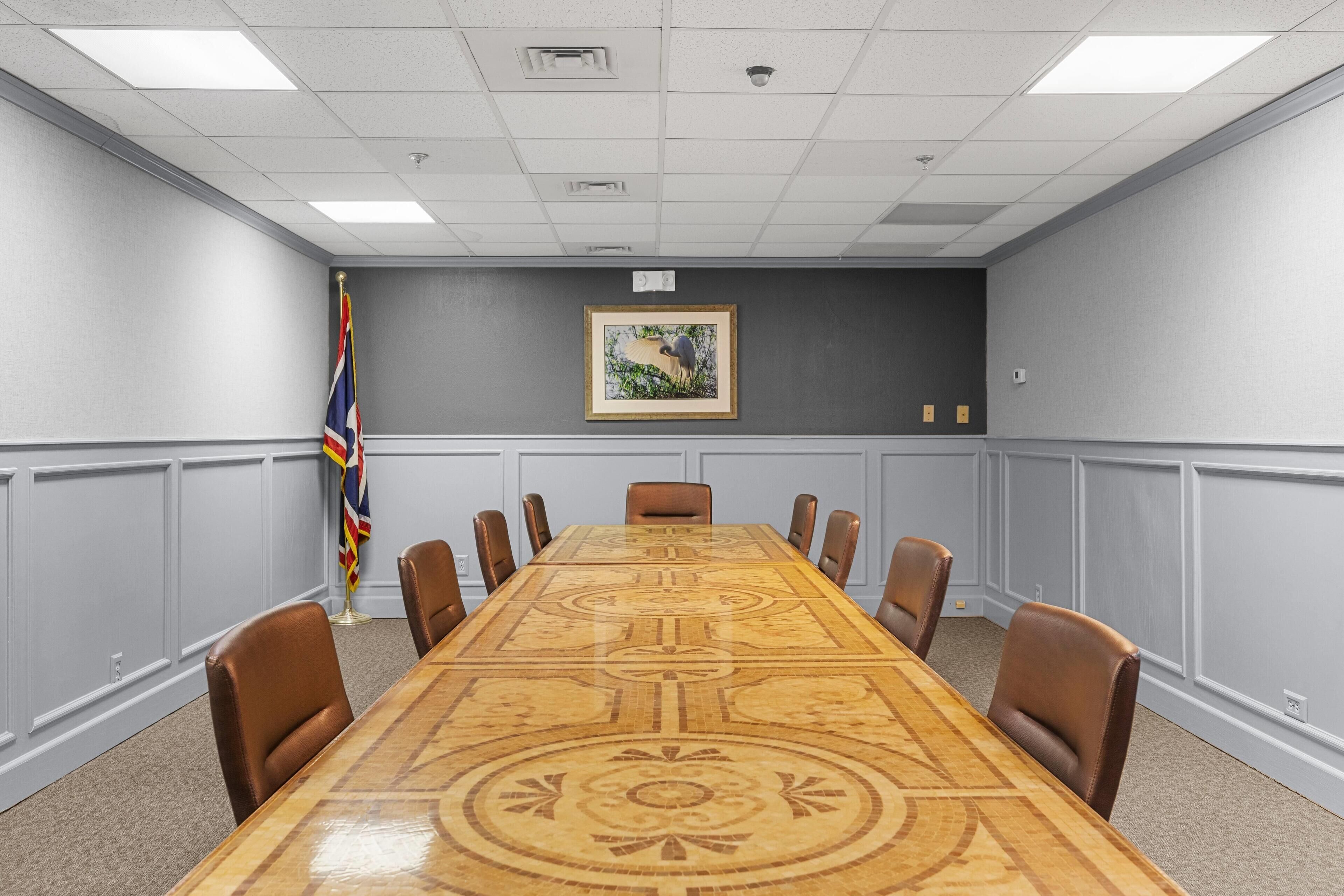 Una sala de conferencias con una larga mesa, sillas de cuero marrón, una bandera y un cuadro en la pared.