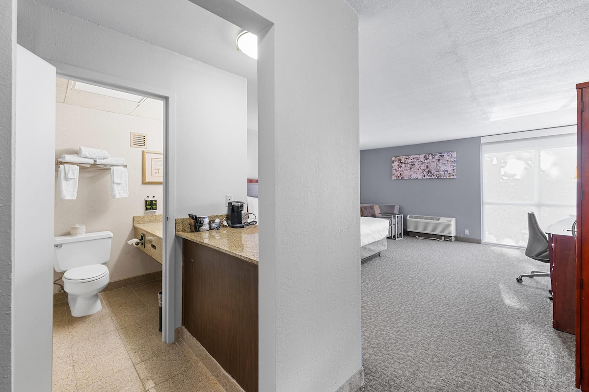 Quarto de hotel com banheiro, cozinha compacta, cama, escrivaninha e uma janela grande.
