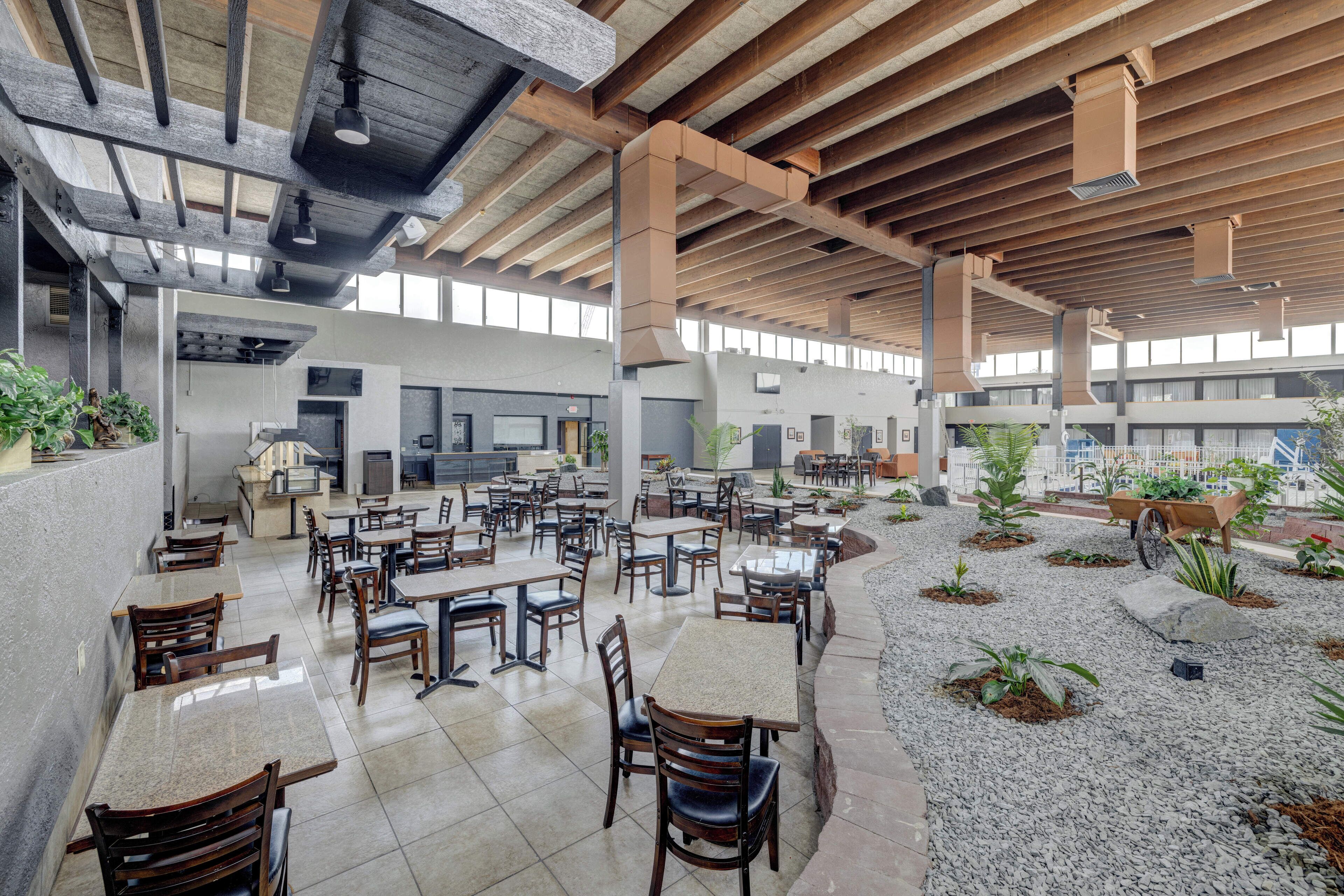 Un amplio comedor interior con varias mesas y sillas, una zona de estar central con un banco, y un pequeño jardín con plantas y una bicicleta.
