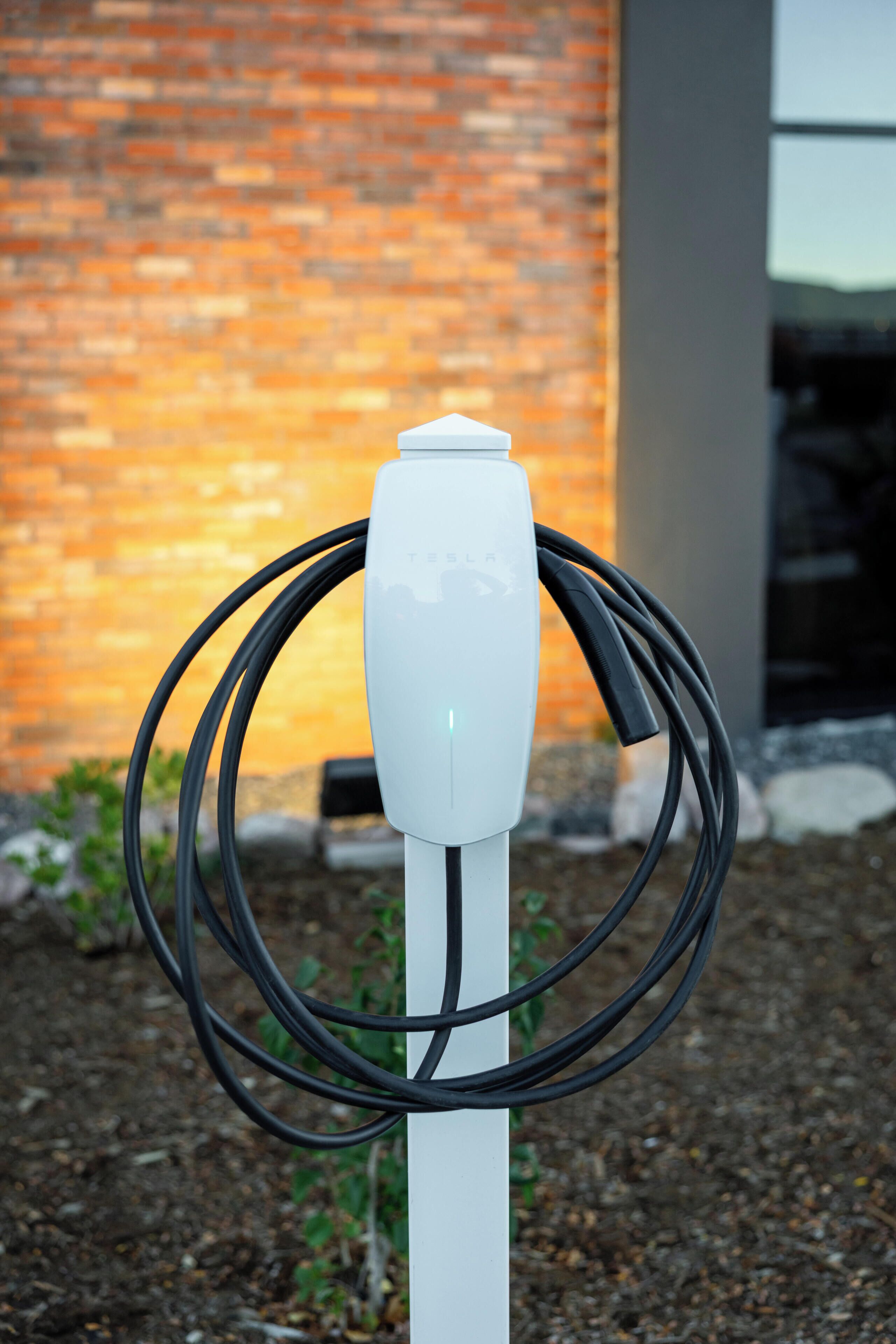 A white electric vehicle charging station with a brick wall and a modern building in the background.
