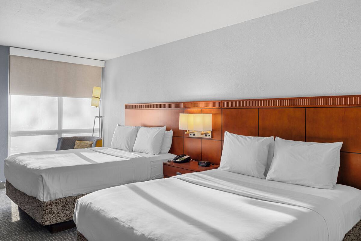 Quarto de hotel com duas camas, uma mesa de cabeceira, uma luminária e uma janela com vista.