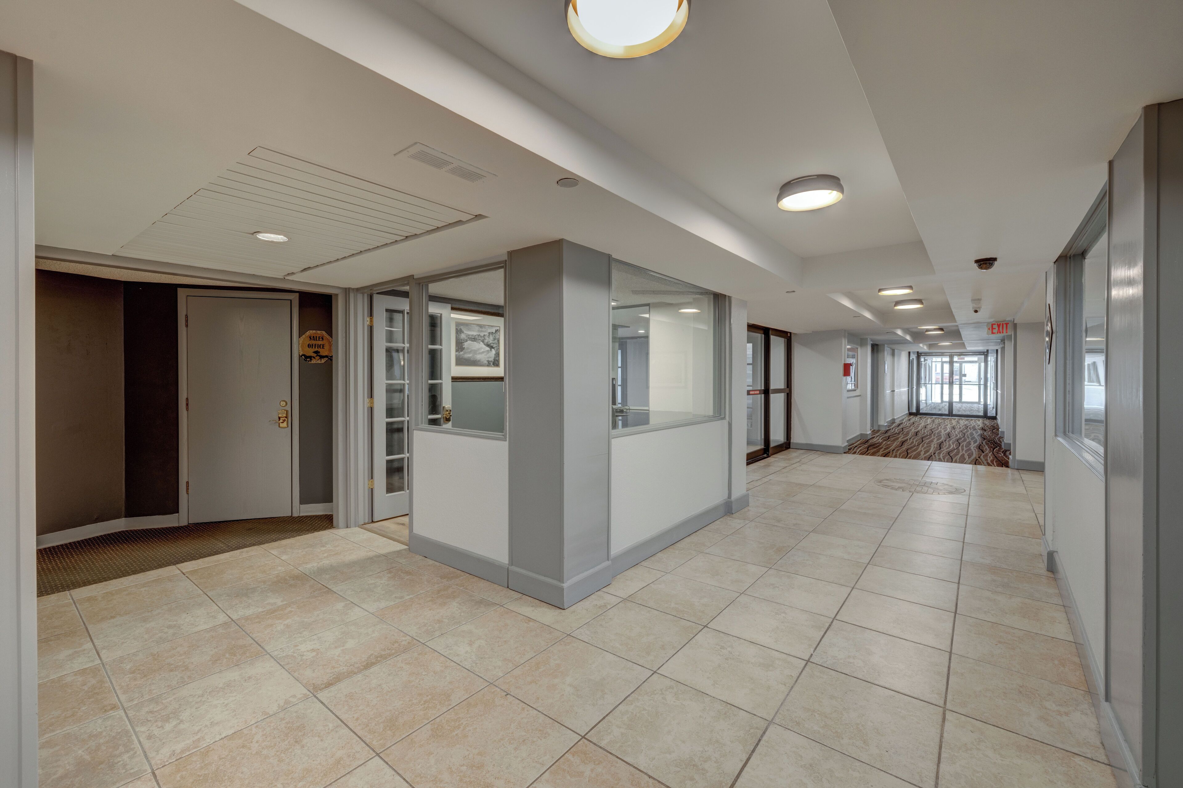 A spacious corridor with tiled flooring, glass partitions, and recessed ceiling lights.
