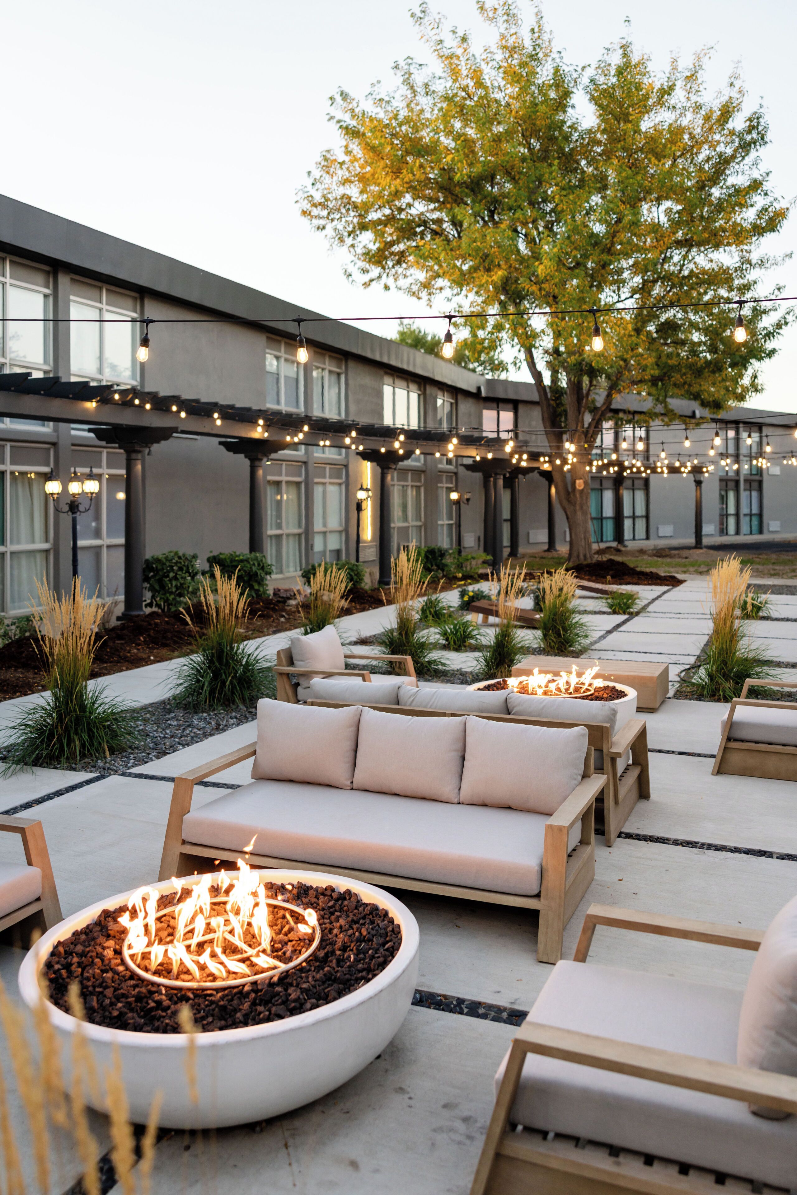 An outdoor seating area with sofas and a fire pit, surrounded by plants and a building with large windows.