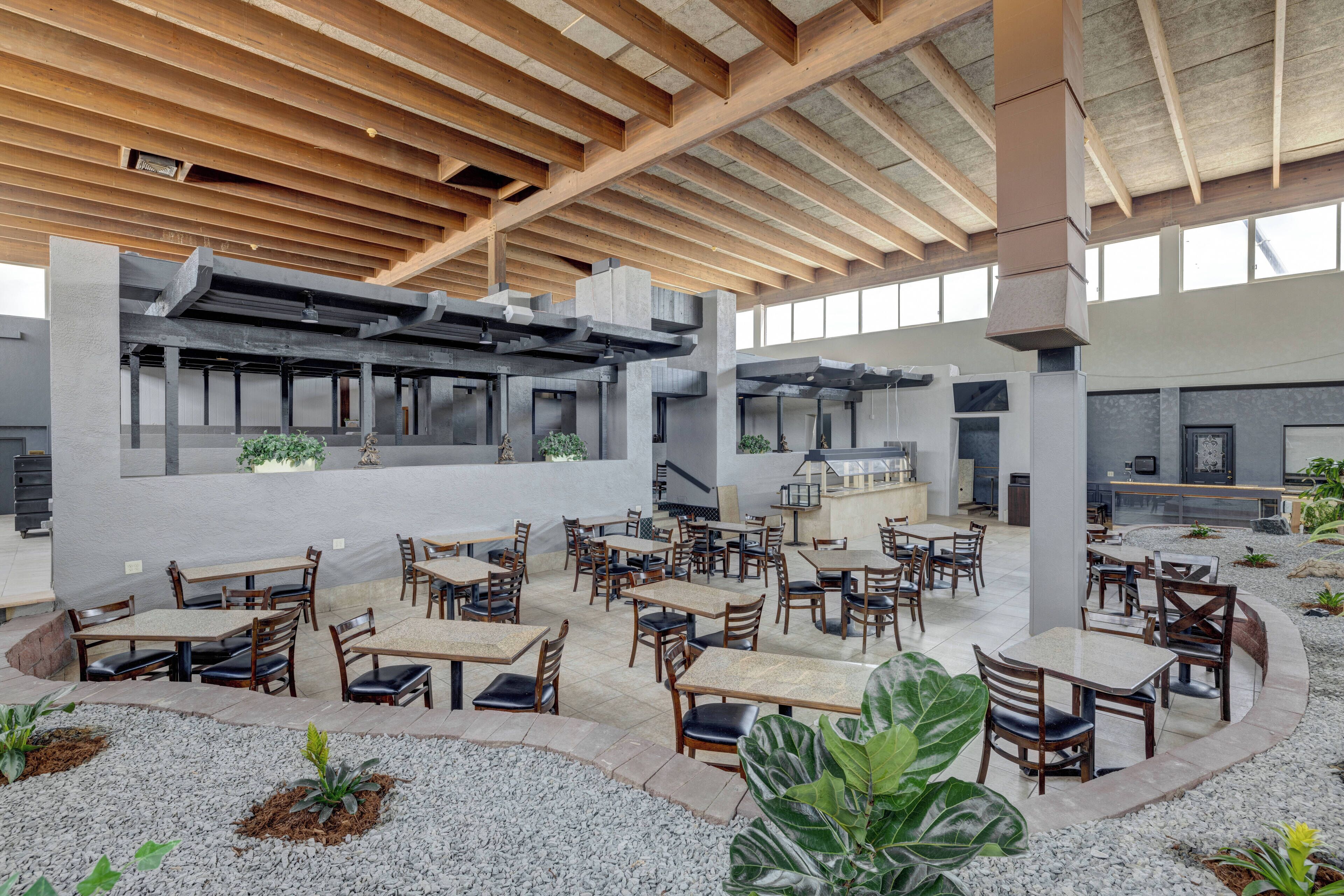 An indoor dining area with wooden tables and chairs, a central kitchen area, and a view of the sky through large windows.