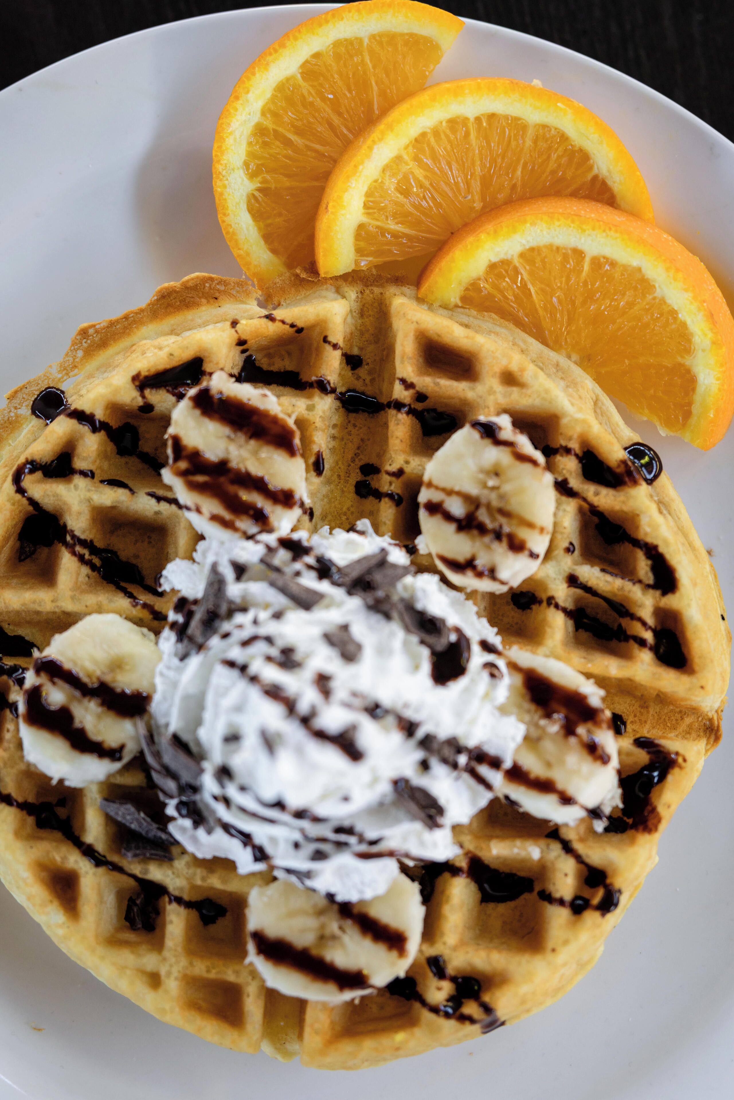 A waffle with whipped cream and banana slices, accompanied by orange slices on a plate.