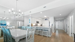 A spacious kitchen with white cabinetry, a central island, and a dining area with a large table and blue chairs. - Salty Breezes (Navarre)