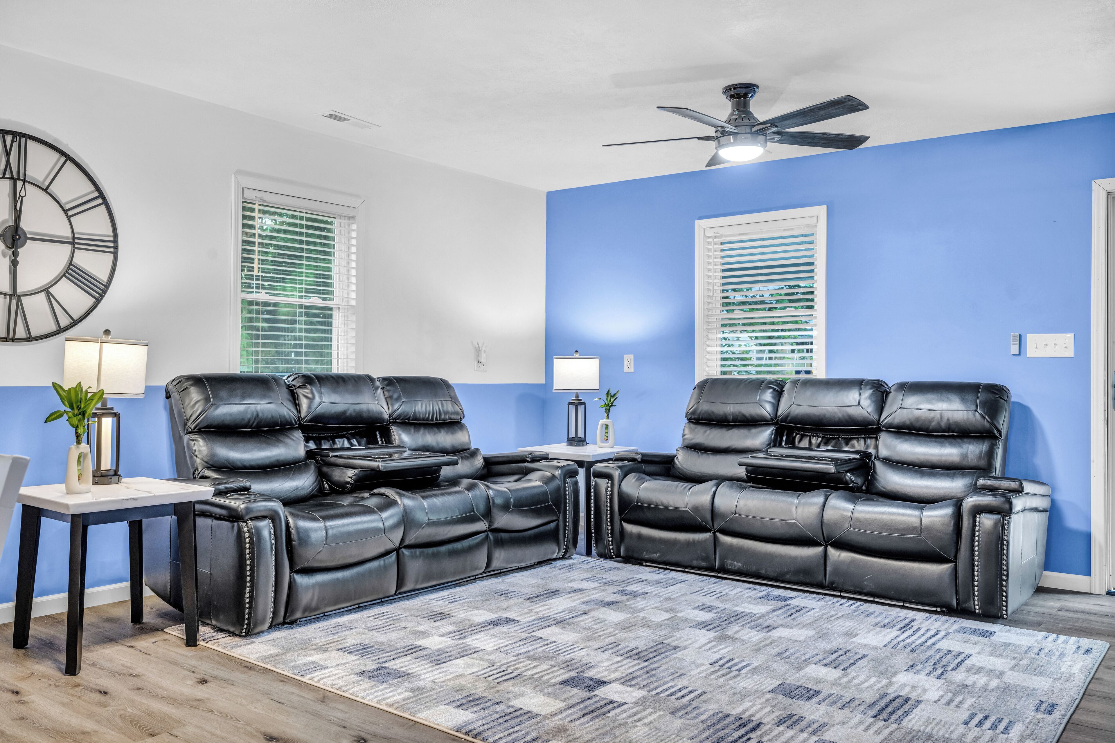 A living room with two black leather sofas, a coffee table, a side table with a lamp, a large wall clock, and a ceiling fan.