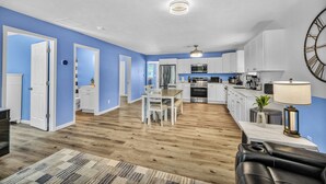 A modern kitchen with white cabinets, a dining area with a table and chairs, and a living room with a leather sofa and a patterned rug.
