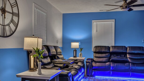 A living room with blue walls, a ceiling fan, a large clock, a massage chair, a massage table, and a patterned rug.