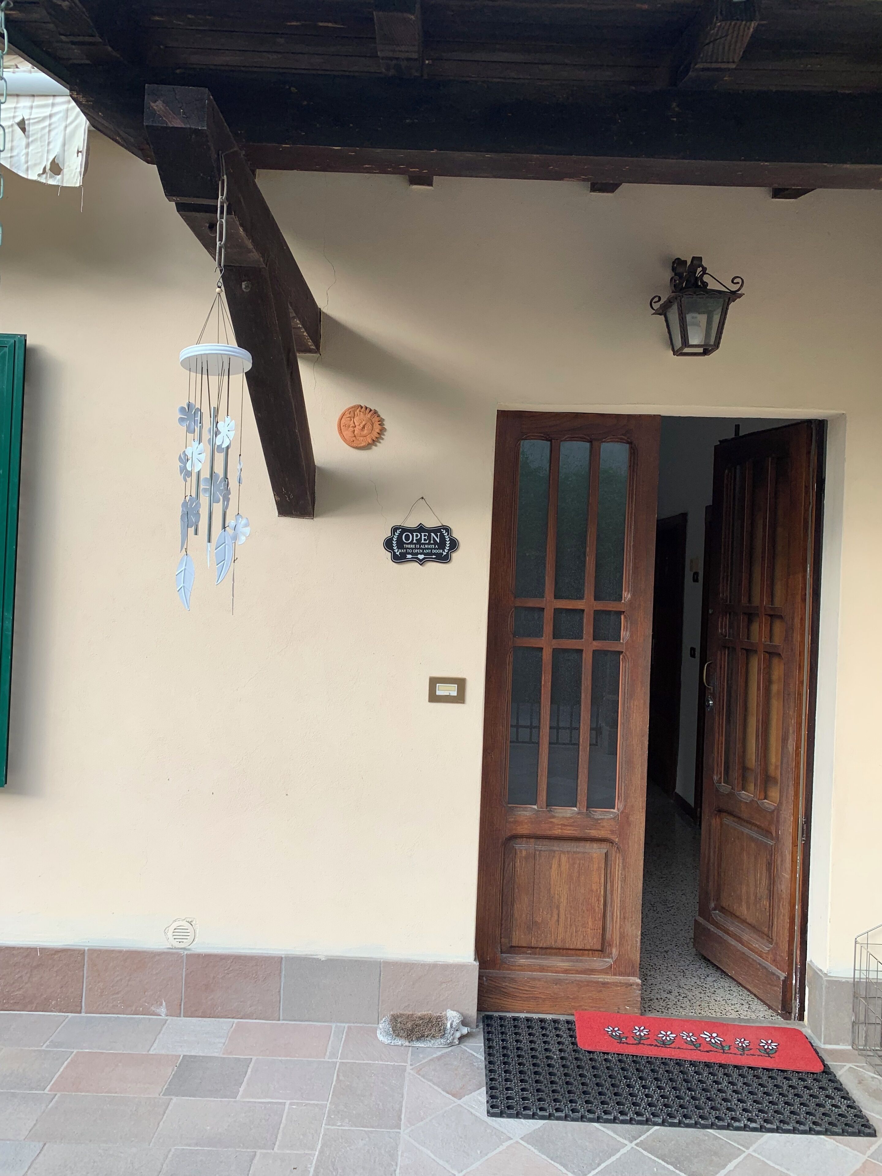 Una porta in legno con un pannello di vetro, una targa fissata al muro, un campanello a vento appeso e un tappetino d'ingresso con motivo floreale.