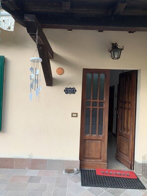 A wooden door with a glass panel, a wall-mounted sign, a hanging wind chime, and a doormat with a floral design. - Apartment, private garden, bright, quiet, pets allowed. (Camporgiano)