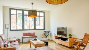 Un salon moderne avec un canapé en cuir, une table basse en bois, un meuble pour la télévision et une grande fenêtre donnant sur un immeuble en briques.
