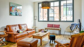 Un salon spacieux avec un canapé en cuir marron, une table basse en bois et un pouf noir. On y trouve un banc en osier avec des coussins à rayures rouges et blanches, une chaise en osier et une grande fenêtre donnant sur un bâtiment en briques.