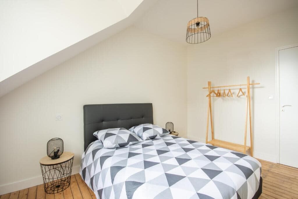 Ein Schlafzimmer mit schrägem Dach, einem Bett mit geometrischem Muster auf der Bettdecke, einem Nachttisch mit Lampe und einem Holztrockner an der Wand.