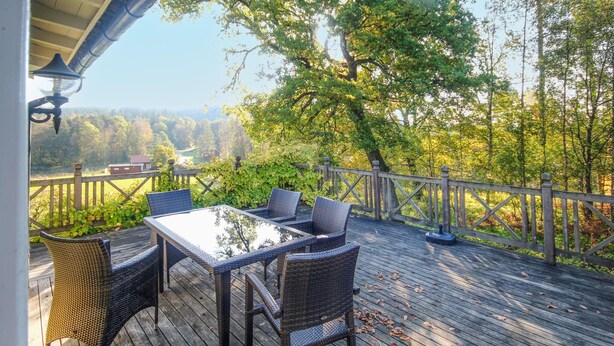 Eine Holzterrasse mit Korbmöbeln und einem Glastisch, mit Blick auf eine bewaldete Gegend.