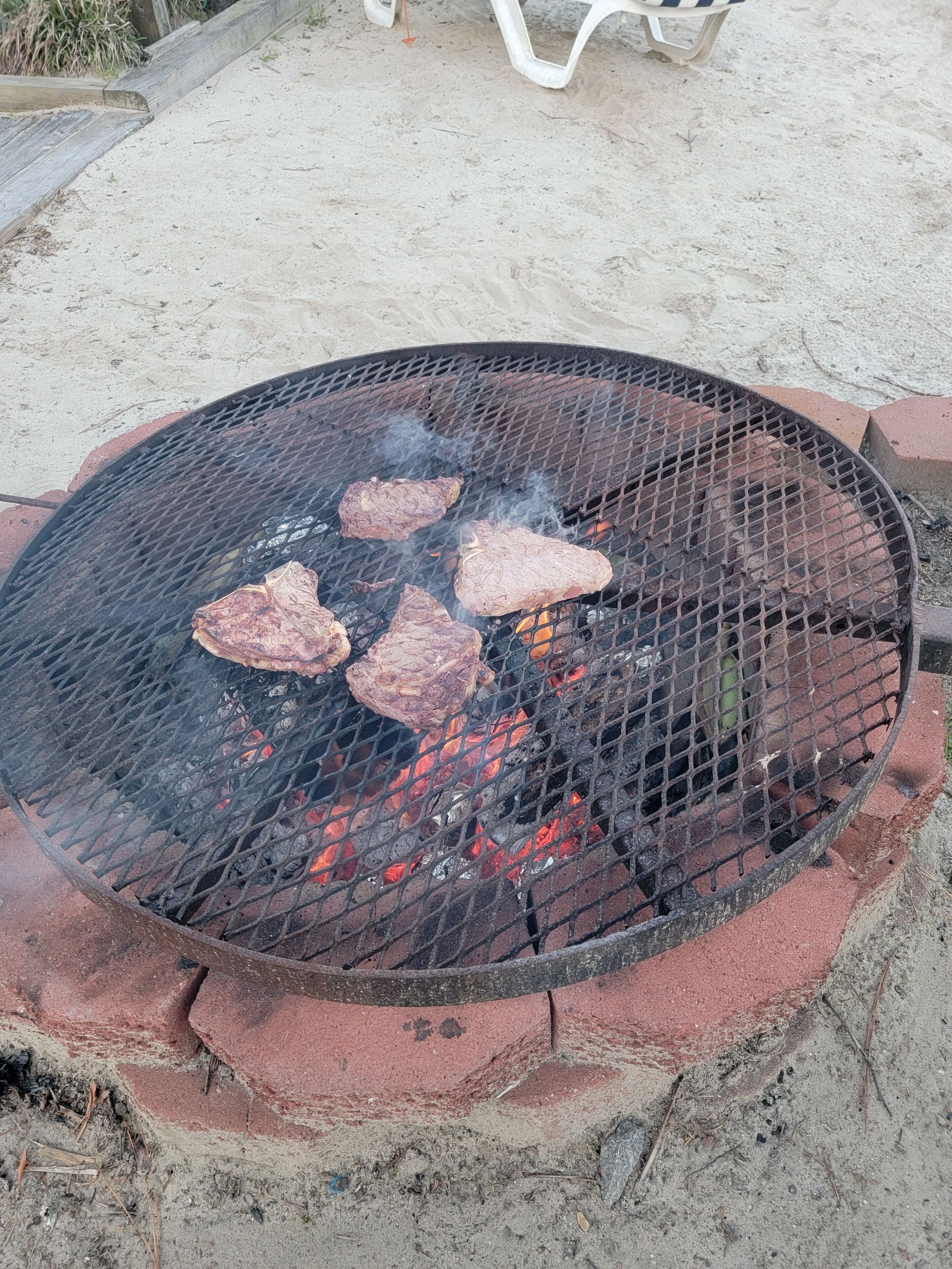 Una parrilla al aire libre, redonda, con cuatro piezas de carne asándose sobre fuego vivo.
