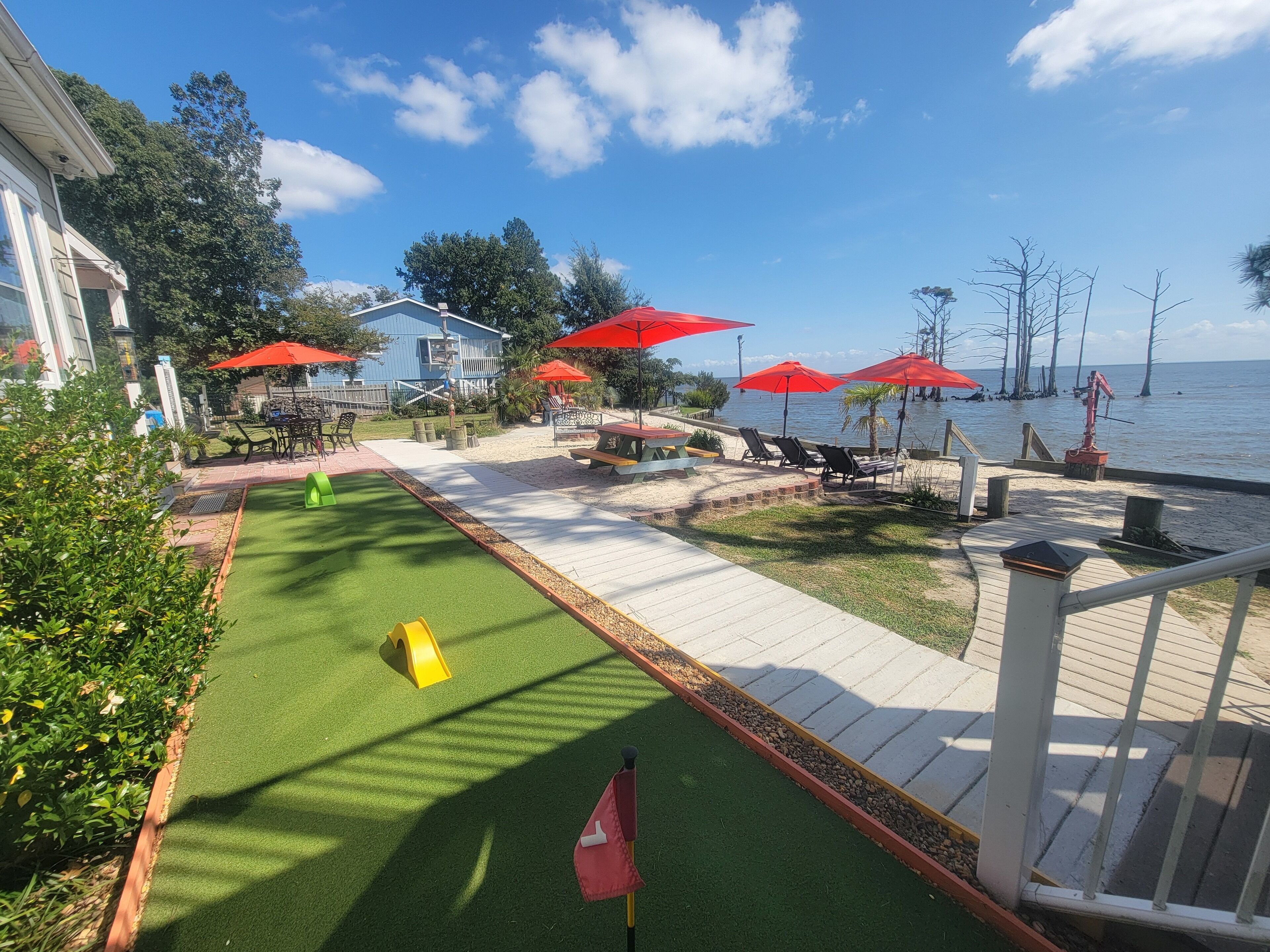 A deck area with a green artificial turf, outdoor seating, red umbrellas, and a view of the water.