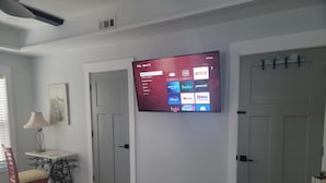 A hotel room with a mounted TV displaying various streaming services, a door, a small table with a lamp, and a chair. - Edgewater Pirates Den. Water Front 2nd story apt.  1 bedroom, bath & kitchen.
 (Hertford)