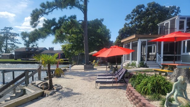 A sandy beach area with lounge chairs and red umbrellas, a wooden deck, and a house with a porch. - Edgewater Pirates Den. Water Front 2nd story apt.  1 bedroom, bath & kitchen.
 (Hertford)