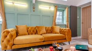 Ein gemütliches Wohnzimmer mit einem senfgelben Sofa, einem Holztisch mit Snacks und Getränken und einem Ausblick ins Freie durch ein Fenster.
