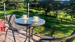A round table with two white cups and saucers, surrounded by metal chairs, set on a balcony with a view of a vineyard and mountains.