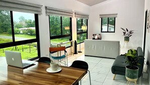 A modern living room with a wooden dining table, a sofa, and a large window offering a view of the outdoors.