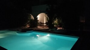 Une piscine à l’eau d’un bleu cristallin, entourée d’arbres et bordée d’une terrasse avec des tables et des chaises.
