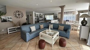 A spacious living room with a blue sectional sofa, a white coffee table, and two leather ottomans. A large wall clock and a modern kitchen are visible in the background. - Villa Dunisia - Leon (LEON)