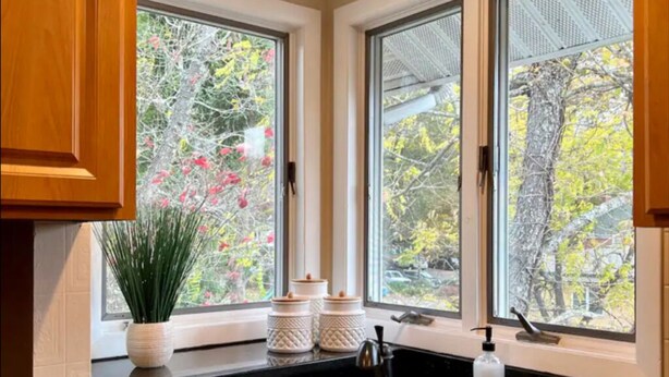 Una cocina con encimera negra, armarios de madera, pileta de acero inoxidable y ventanas amplias con vistas a árboles y un edificio.