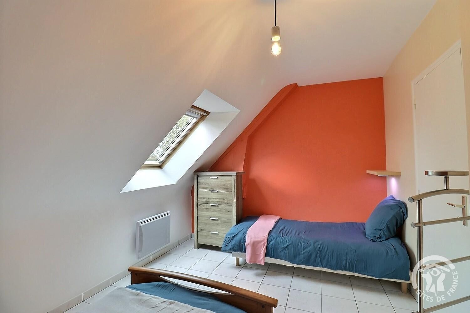 Un dormitorio con techo inclinado, un tragaluz, cama con ropa de cama azul, cómoda de madera y lámpara colgante.