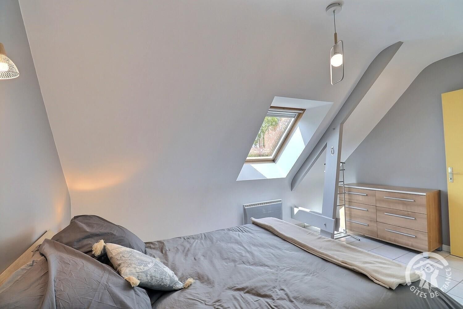 A bedroom with a sloped ceiling, a skylight, a bed with pillows, a bedside table, and a wardrobe.