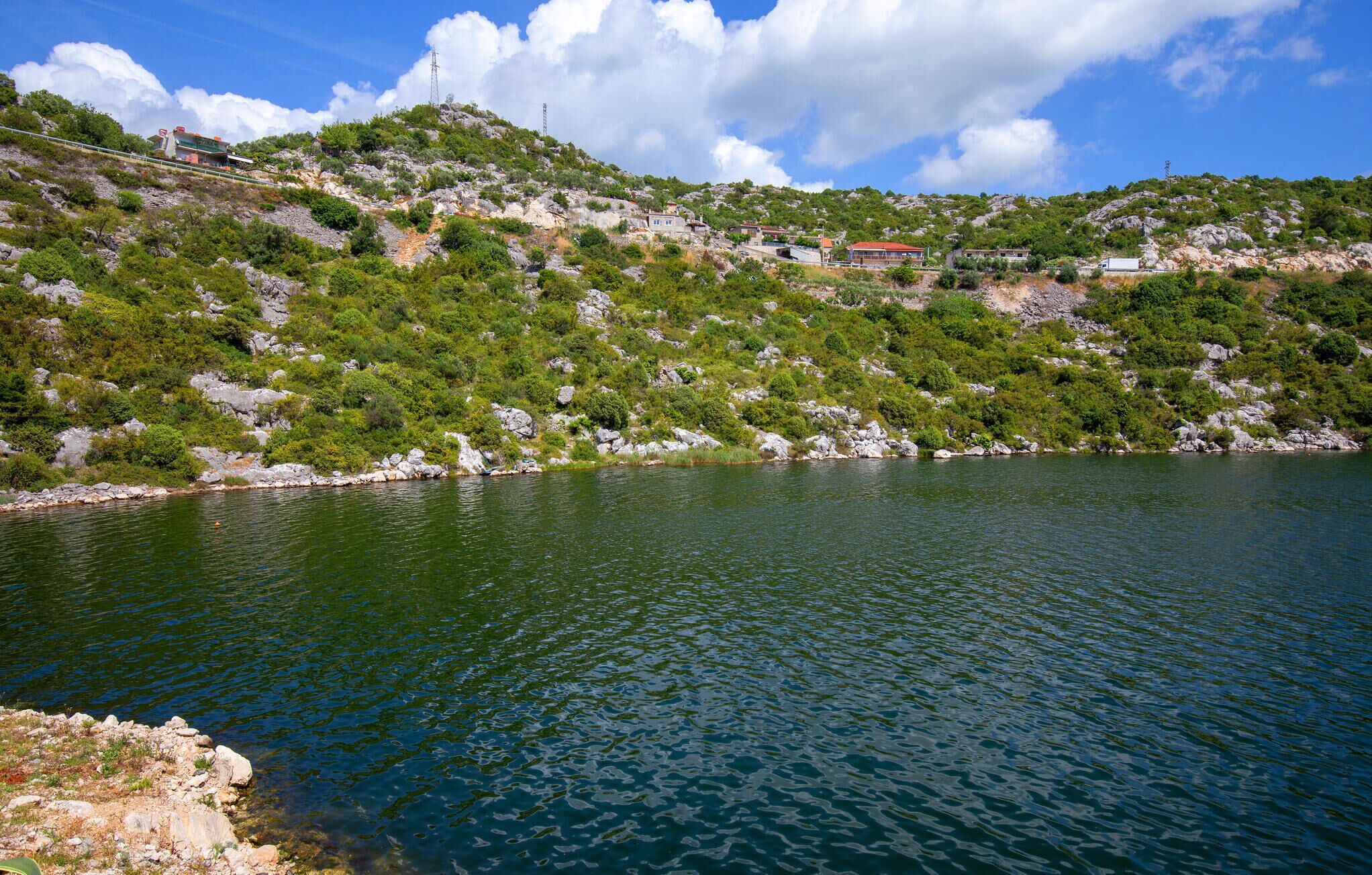 Un lac entouré de collines rocheuses, avec une végétation clairsemée et quelques bâtiments visibles au loin.